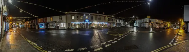 A night panorama of Great Dunmow’s Market Place and High Street under strings of Christmas lights: rows of lit shopfronts and the Saracen’s Head hotel, decorated with blue lights and a star, around a wet, empty junction