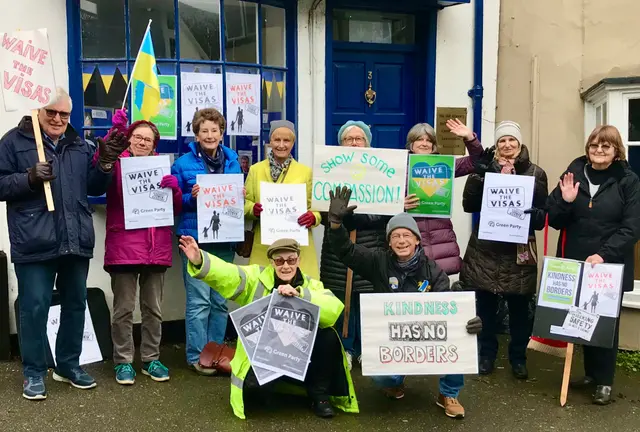 Ten people, smiling and waving, with a Ukrainian flag and placards reading “Waive the visas” and “Kindness has no borders”, outside a building with a blue door.